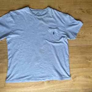 Grå t-shirt från Polo Ralph Lauren - Säljer en klassisk grå t-shirt från Polo Ralph Lauren med en liten bröstficka och broderad logga. Perfekt för en avslappnad stil. T-shirten är kortärmad och tillverkad i mjuk bomull.