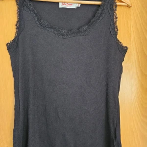 Svart ärmlös topp från John Baner - Säljer en stilren svart ärmlös topp från John Baner. Toppen har en ribbad struktur och är dekorerad med spetsdetaljer runt halsringningen och ärmöppningarna. Perfekt för en elegant och bekväm look.  Sparsamt använd. Djur och -rökfritt hem.