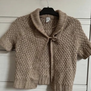Beige stickad kofta från H&M kortärmad - Säljer en mysig beige stickad kofta från H&M med korta ärmar och en stor knapp framtill. Koftan har ett fint stickat mönster och en bred krage. Perfekt för lager-på-lager-stil! 🧶