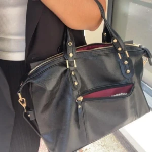 Väska - Snygg svart väska i skinn med gulddetaljer och dragkedja. Väskan har en praktisk ytterficka och röd insida som ger en fin kontrast. Perfekt för att bära med sig det nödvändigaste med stil. Lånade bilder!  Aldrig använd. Köpt för 200kr