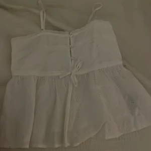 Vit ärmlös topp från Gina Tricot - Söt vit ärmlös topp från Gina Tricot med broderade detaljer och knytband i midjan. Toppen har smala axelband och en luftig passform, perfekt för varma dagar. Den har en fin struktur med små hålmönster 💞