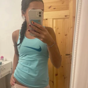 Ljusblå linnetopp från Nike - Säljer en ljusblå linnetopp från Nike med det klassiska swoosh-loggan i en mörkare blå nyans. Toppen har en tight passform och är ärmlös, perfekt för träning eller varma sommardagar. Linnet är i storlek M i barn storlek men sitter som en xs vuxen