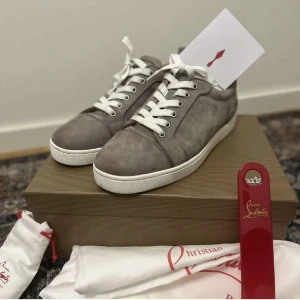 Grå sneakers från Christian Louboutin - Snygga grå sneakers i mocka från Christian Louboutin med den ikoniska röda sulan. Skorna har vita snören och en vit sula som ger en stilren look. Perfekta för dig som vill ha en touch av lyx i vardagen.