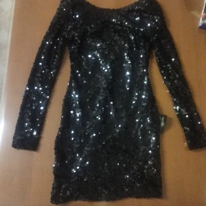 Svart paljettklänning - Snygg svart klänning täckt med glittriga paljetter. Den har en åtsittande passform och långa ärmar, perfekt för en festlig kväll. Klänningen är kort och har en rund halsringning.