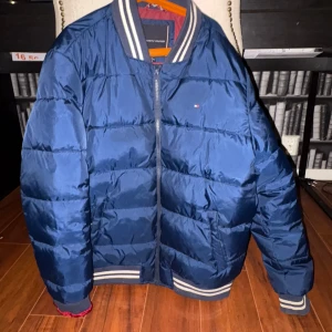 Blå pufferjacka från Tommy Hilfiger - Snygg blå pufferjacka från Tommy Hilfiger med dragkedja och ribbade muddar. Jackan har ett stort tryck med märkesnamnet på ryggen och en klassisk design med långa ärmar. Perfekt för kyligare dagar.