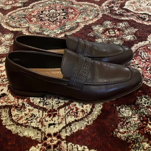 Bruna loafers i skinn - Snygga bruna loafers i skinn med flätad detalj över vristen. Perfekta för en stilren look. Storlek 44.