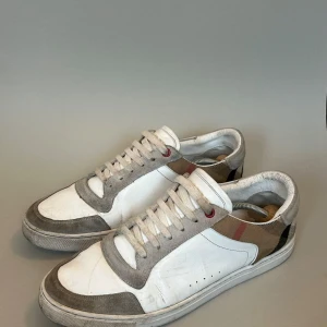 Vita sneakers från Burberry - Snygga vita sneakers från Burberry med gråa detaljer och klassiskt rutigt mönster på insidan. Skorna har snörning och en stilren design som passar perfekt för en avslappnad look.