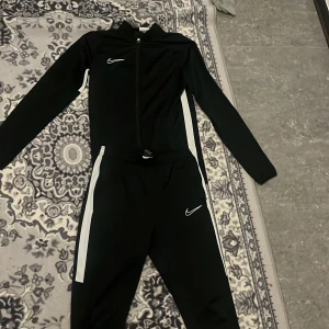 Svart träningsdräkt från Nike - Snygg svart träningsdräkt från Nike med vita detaljer. Jackan har dragkedja och byxorna har elastisk midja. Perfekt för sport och träning.