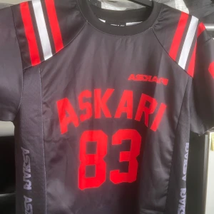 Askari T-shirt  - Svart t-shirt med röda detaljer och texten "ASKARI 83" på framsidan och baksidan. T-shirten har en rund halsringning och korta ärmar.