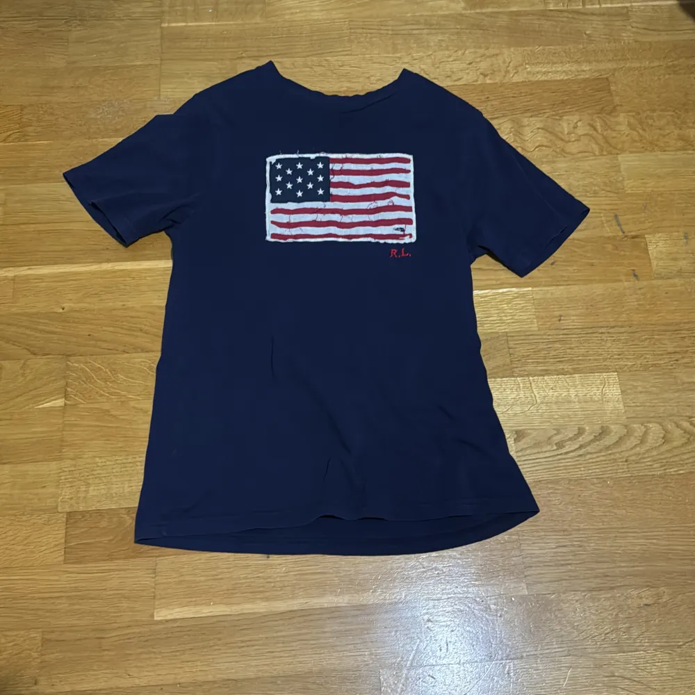 Säljer en snygg mörkblå t-shirt från Ralph Lauren med en cool amerikansk flagga på framsidan. T-shirten är i bomull och har korta ärmar. Perfekt för en avslappnad stil eller en somrig dag. Flaggan har en vintage-look med L barn är s i vuxen tror ja . T-shirts.