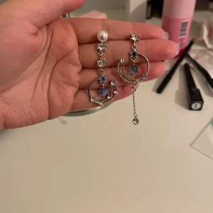 Silverfärgade örhängen med pärlor och stjärndetaljer - Säljer ett par unika och eleganta silverfärgade örhängen. De har en vacker design med pärlor och små stjärnor i blått och silver. Ena örhänget har en längre kedja som ger en asymmetrisk look. Perfekta för en fest eller en speciell kväll!