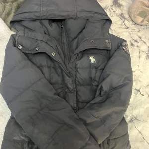 Svart dunjacka från Abercrombie & Fitch - Säljer en snygg och varm svart dunjacka från Abercrombie & Fitch. Jackan har en huva och dragkedja framtill, samt ett stilrent märke på ärmen. Perfekt för kalla vinterdagar! 🖤