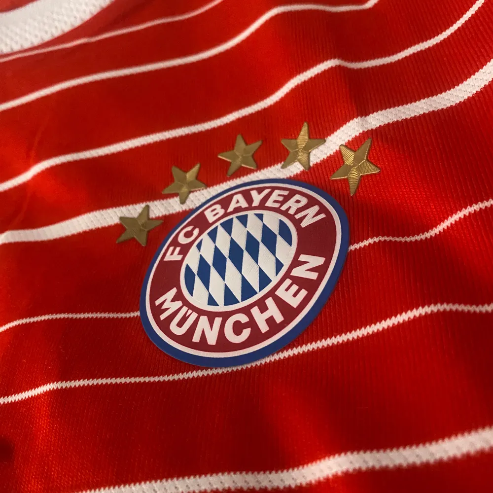 Säljer en snygg röd FC Bayern München fotbollströja med vita ränder. Tröjan har korta ärmar och är i bra skick. Den har klubbens emblem på bröstet och Adidas-loggan. På baksidan står det 'FC Bayern München' och nummer 10 med namnet 'Sané'. Perfekt för alla Bayern-fans!. T-shirts.