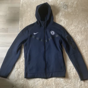 Nike tech Chelsea - Säljer en snygg mörkblå hoodie från Nike med Chelsea FC-logga på bröstet. Den har en dragkedja framtill och är långärmad med en skön huva. Perfekt för fotbollsfans som vill hålla sig varma och stiliga. Ny pris är 1699kr mitt pris 400kr