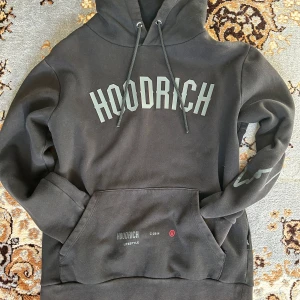 Svart hoodie från Hoodrich - Säljer en svart hoodie från Hoodrich med tryck på framsidan och ärmarna. Den har en stor ficka fram och en justerbar huva med dragsko. Perfekt för en casual look eller att mysa i hemma. Superbekväm och stilren!