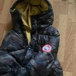 Camouflage pufferjacka från Canada Goose - Säljer en varm pufferjacka från Canada Goose i ett  camouflage-mönster. Jackan har en mysig huva med gul insida. Perfekt för kalla vinterdagar! Har ett hål på framsidan därav låga priset