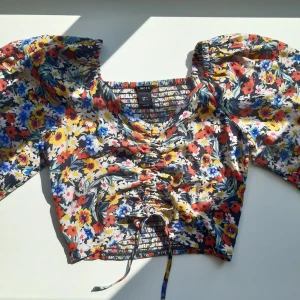 Blommig blus - Vacker blommig blus i ett mjukt skönt tyg. Har långa ärmar, stretch på baksidan för bättre passform och den knyts framtill så att man själv kan justera den så som man vill. Aldig använd - perfekt skick! Säljer en likadan i en vacker grå-grön färg! 