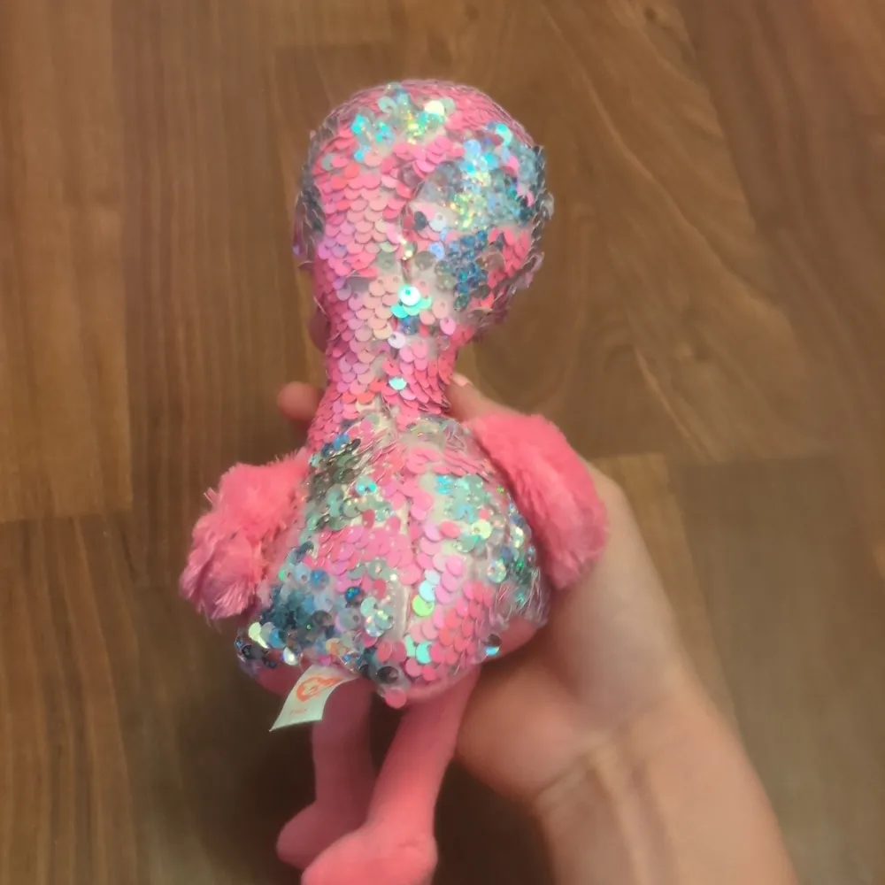 Supergullig rosa flamingo gosedjur täckt med glittriga paljetter som skiftar i färg. Den har stora svarta ögon och fluffiga rosa vingar. Perfekt för att lysa upp vilket rum som helst eller som en söt present!. Asusteet.