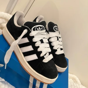 Svarta Adidas Campus sneakers - Säljer ett par snygga svarta Adidas Campus sneakers i mocka. De har de klassiska vita tre ränderna på sidan och en vit sula. Skorna är i nyskick och kommer i originalkartong. Perfekta för både vardag och fest!