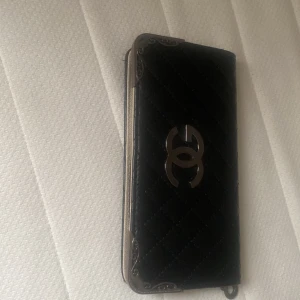 Svart quiltad plånbok - Säljer en elegant svart quiltad plånbok med metallhörn och ett stilrent märke på framsidan. Den har en dragkedja och är perfekt för att hålla dina kort och kontanter säkra. Passar både till vardags och fest!