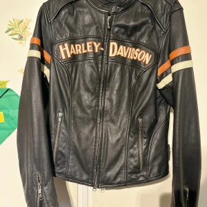 Svart skinnjacka från Harley-Davidson - Säljer en svart skinnjacka från Harley-Davidson med coola detaljer. Jackan har långa ärmar och dragkedja framtill. Den har också snygga orange och vita ränder på ärmarna och ett stort Harley-Davidson-märke på ryggen. Köpte i New York. Passa storlek s/m