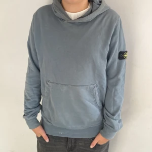 Blå hoodie från Stone Island - Säljer en snygg blå hoodie från Stone Island. Den har en klassisk känguruficka och en bekväm huva. Perfekt för en avslappnad stil och passar till alla tillfällen. Märkesloggan sitter på ärmen för en extra touch av stil. Skön och enkel att matcha med jeans eller joggers.