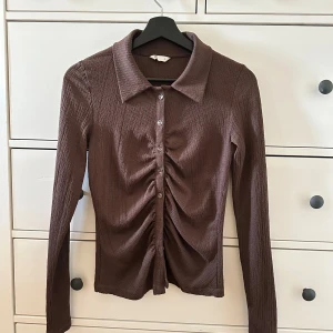 Brun ribbad skjorta med knappar - Säljer en  brun ribbad skjorta med långa ärmar och knappar framtill. Skjortan har en cool rynkad detalj i mitten som ger den en unik look. 