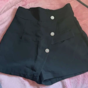 Svarta shorts - Säljer ett par stilrena svarta shorts från New Collection. De är högmidjade med tre snygga knappar framtill och en dragkedja bak. Perfekta för både vardag och fest!