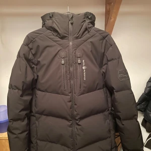 Svart dunjacka från Sail Racing - Säljer en svart dunjacka från Sail Racing, perfekt för kalla vinterdagar. Jackan har en stilren design med dragkedja och flera praktiska fickor. Den är långärmad och har en justerbar huva för extra skydd mot väder och vind. Den är i fint använt skick