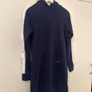 Mörkblå hoodieklänning från GANT - Säljer en snygg mörkblå hoodieklänning från GANT. Perfekt för en avslappnad stil och superbekväm att ha på sig. Stor ficka framtill för extra mysfaktor. Passar perfekt till höst och vår!