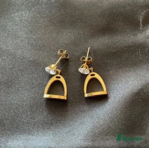 Guldörhängen med stigbygel-design - Snygga guldörhängen i form av stigbyglar, perfekta för att ge en unik touch till din outfit. De har en elegant och stilren design med en liten glittrig detalj upptill. Passar både till vardags och festliga tillfällen. Ett måste för den som älskar hästtema!