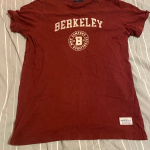 Röd t-shirt från Berkeley - Säljer en snygg röd t-shirt från Berkeley i storlek M. Den har ett tryck med texten 'Berkeley' och en rund logga på framsidan. Perfekt för en avslappnad stil. T-shirten är i bomull och har korta ärmar. Passar bra till både jeans och shorts!