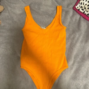 Orange ribbad bodysuit - Säljer en snygg orange ribbad bodysuit i mycket bra skick. Den är ärmlös och har en djup rundad halsringning. Perfekt för sommardagar eller som ett statement-plagg till fest. Materialet är stretchigt och bekvämt.