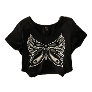 Svart croppad t-shirt med fjärilsmotiv - Snygg svart croppad t-shirt från Shein med ett coolt fjärilsmotiv i silver på framsidan. Perfekt för en avslappnad stil eller en utekväll. T-shirten har en v-ringad hals och korta ärmar.