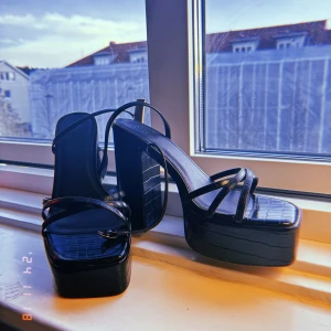 Svarta platåsandaler med klack - Säljer ett par snygga svarta platåsandaler med hög klack och remmar.  STORLEK 39 Perfekta för en utekväll eller fest! De har en cool krokodilpräglad yta som ger en extra touch av stil. Endast testade hemma. Passar perfekt till både klänning och jeans.