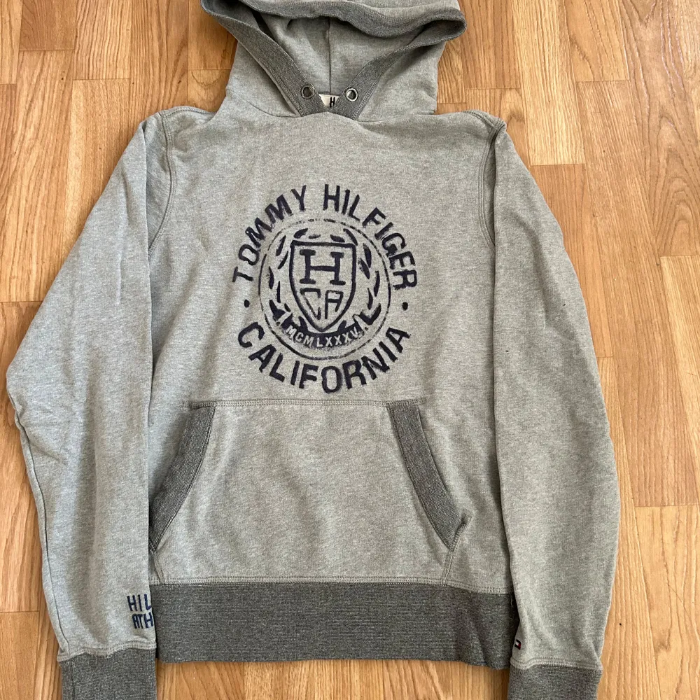 Säljer en snygg grå hoodie från Tommy Hilfiger med tryck 'California' och logga på framsidan. Den har en känguruficka och ribbade muddar. Perfekt för en avslappnad stil eller kyliga dagar. Passar både till vardags och chillkvällar. 🕶️. Tröjor & Koftor.