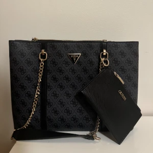 Tote väska guess - Snygg black multi tote väska från Guess med kedjedetaljer och matchande plånbok. Väskan finns att hämta i Salem eller fraktas.