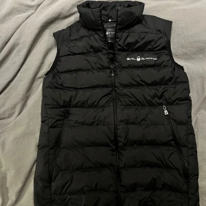 Svart dunväst från Sail Racing - Säljer en svart dunväst från Sail Racing i mycket bra skick. Västen har en dragkedja framtill och två praktiska fickor med dragkedjor. Perfekt för höst och vår när du vill hålla dig varm utan att bära en jacka. Den quiltade designen ger en stilren look.