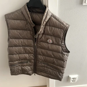 Brun dunväst från Moncler  - Säljer en snygg brun dunväst från Moncler i mycket bra skick. Västen har en dragkedja framtill och två praktiska fickor med dragkedjor. Perfekt för höst och vår när du vill hålla dig varm men ändå se stilren ut. Passar både till vardags och lite finare tillfällen. Enda flawen som finns är dragkedjan som är något sne i slutet men inget som påverkar funktionen o är lätt fixat. Priset är självklart rörligt 