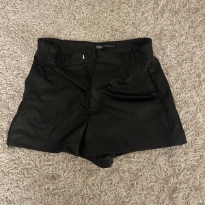 Svarta shorts i skinn från Zara - Säljer ett par snygga svarta skinnshorts från Zara. De är midwaist och har en stilren design med dragkedja fram. Perfekta för en cool look på festen eller en utekväll. Passar bra till både sommar och höst. Nästan oanvända🖤