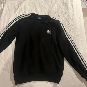 Svart sweatshirt från Adidas - Säljer en klassisk svart sweatshirt från Adidas med de ikoniska tre ränderna längs ärmarna. Tröjan har en normal passform och är perfekt för både vardag och träning. Adidas-loggan är tryckt på bröstet. Superbekväm och stilren! Obs! Storleken står som large, men tycker inte den passar large mer till en smal!