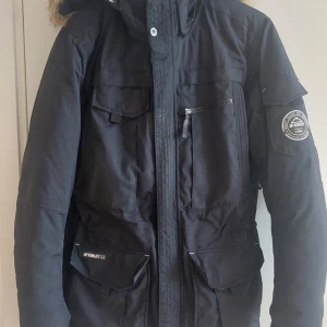 Svart vinterjacka med pälsdetalj - Säljer en svart vinterjacka från Geographical Norway i mycket bra skick. Jackan har en mysig pälsdetalj på huvan och flera praktiska fickor. Perfekt för kalla vinterdagar! Den har en dragkedja och knappar för extra skydd mot kylan. Det är en38 men känns definitivt mer som en 40 eller tom 42. 