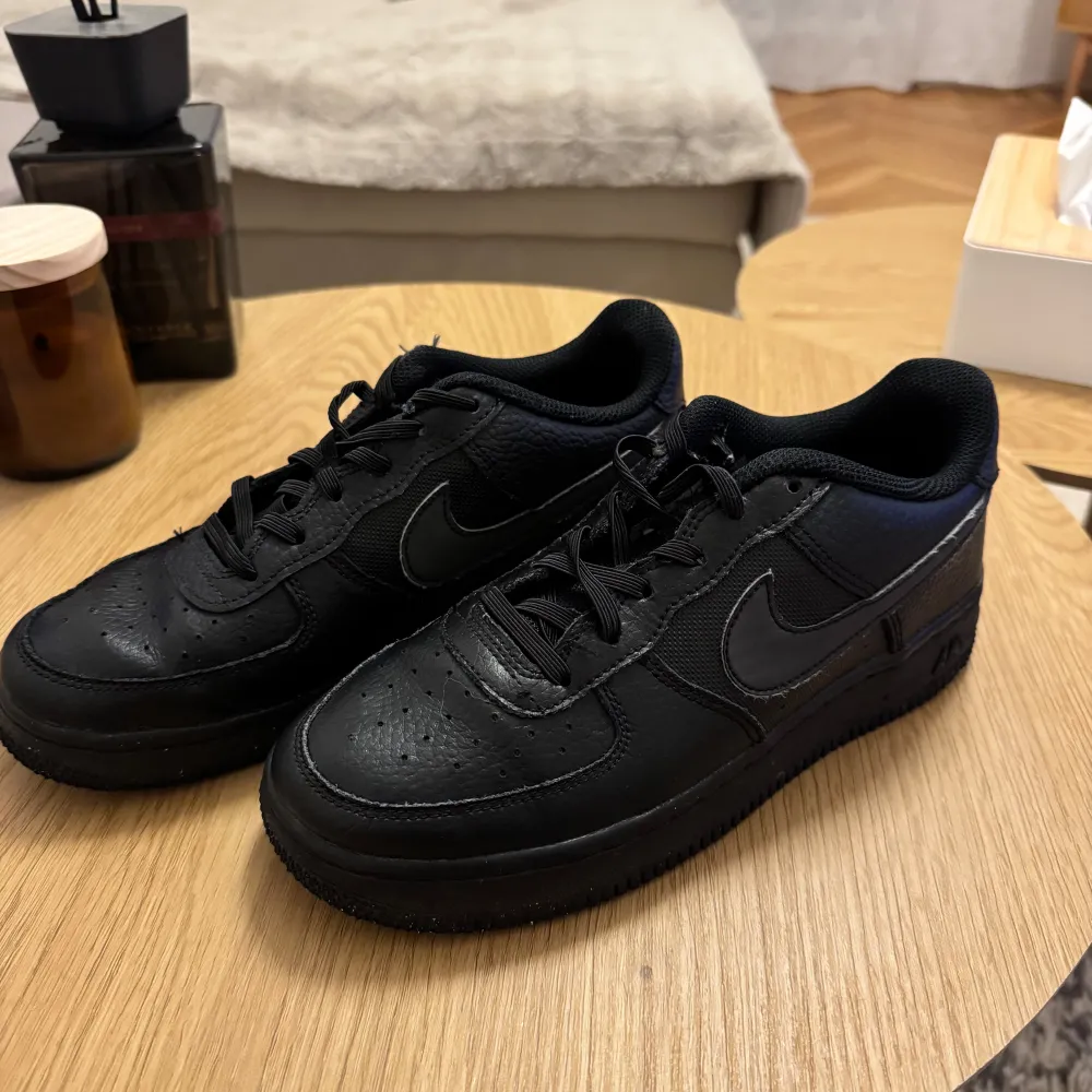 Säljer ett par klassiska svarta Nike Air Force sneakers i mycket bra skick. De är helt svarta med snörning och har den ikoniska Nike-loggan på sidan. Perfekta för både vardag och fest! Storlek och passform är normal.. Skor.