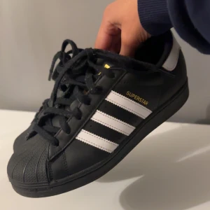 Svarta Adidas Superstar sneakers - Säljer mina sköna och snygga svarta Unisex Adidas Superstars i mycket bra skick. De har de används kanske 3 gånger. Nypris 1500kr. Storlek 37.1/3 men passar även mig som brukar ha allt mellan storlek 37 och 39. Skriv för flera bilder och detaljer! 
