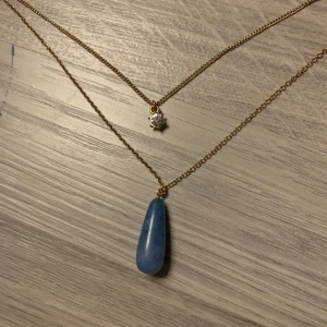 Dubbelhalsband med blå sten och stjärnberlock - Elegant dubbelhalsband med en blå droppformad sten och en liten stjärnberlock. Kedjan är i guldton och justerbar i längd. Ge gärna prisförslag