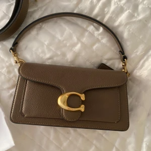 Brun axelväska från Coach - Snygg, brun tabbyväska från Coach i äkta skinn. Använd fåtal gånger och därmed i väldigt fint skick. 