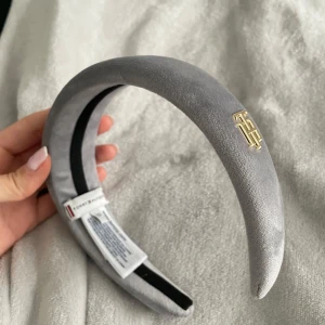 Grått diadem från TommyHilfiger - Elegant grått diadem i sammet med en stilren gulddetalj på sidan. Perfekt för att ge en touch av lyx till din outfit. Diademet har en mjuk och bekväm passform. Helt ny, aldrig använd. Org. pris 400kr.