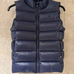 Mörkblå dunväst från Ralph Lauren - Snygg mörkblå dunväst från Ralph Lauren med dragkedja framtill. Västen har en stilren design med diskret logga på bröstet och är perfekt för kyligare dagar. Storleken är Xl i junior storlek så det passar runt Small i vanlig storlek