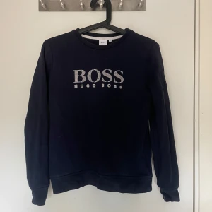Mörkblå tröja från Hugo Boss - Säljer en stilren mörkblå tröja från Hugo Boss med deras logga i silver på bröstet. Tröjan har långa ärmar och en klassisk rund halsringning. Perfekt för en avslappnad men ändå snygg look. Pris är ej hugget i sten