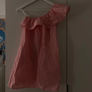 Röd- och vitrandig klänning - Säljer en söt rosa och vitrandig klänning med en offshoulder-design och volangdetalj. Perfekt för sommarens varma dagar. Klänningen har en kort längd och är tillverkad i ett lätt och bekvämt material. Klänningen har fickor men ingenting man ser❤️ köpt i Italien så vet inte märket. Den är stretchig så skulle säga att den passar S-M